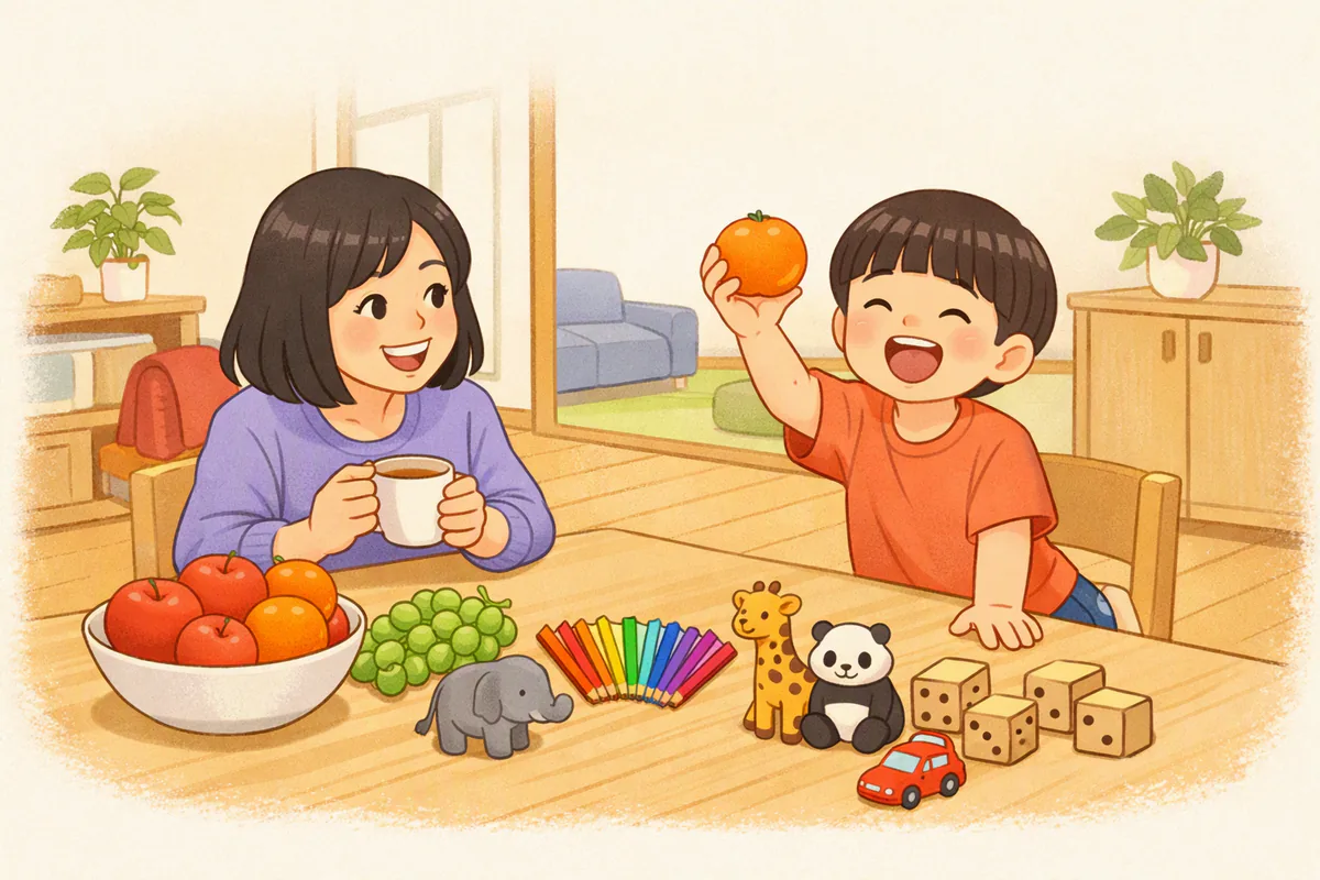 台所のテーブルで、みかんを持ち上げて笑う男の子と、果物・色えんぴつ・動物のおもちゃ、向かいで笑う母親